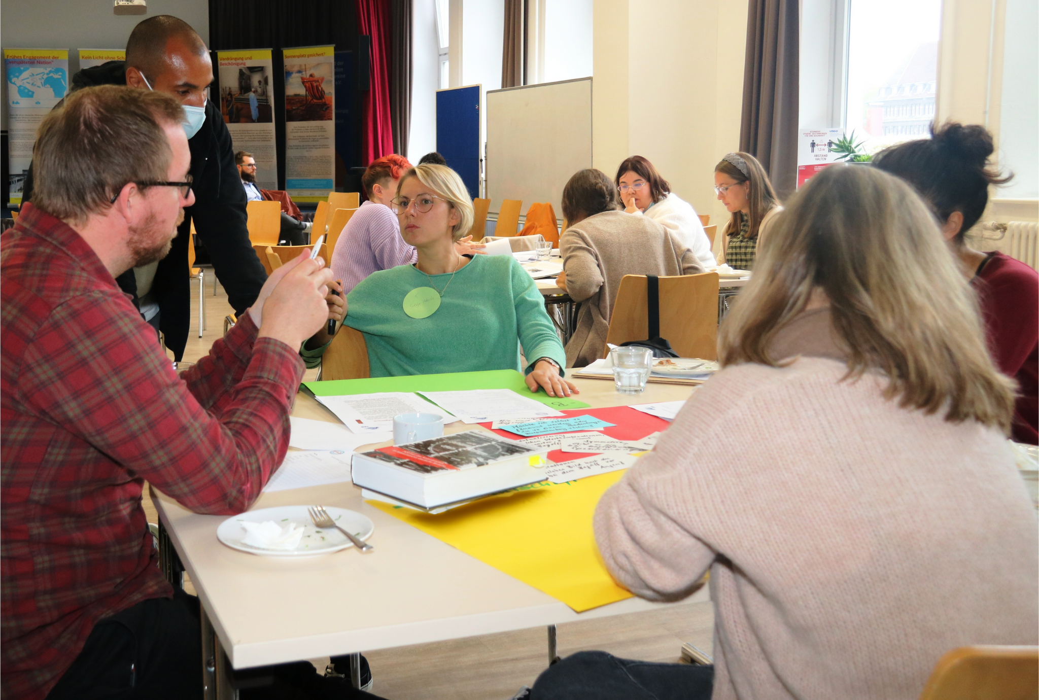 Person mit Maske steht an Tisch mit Teilnehmer:innen